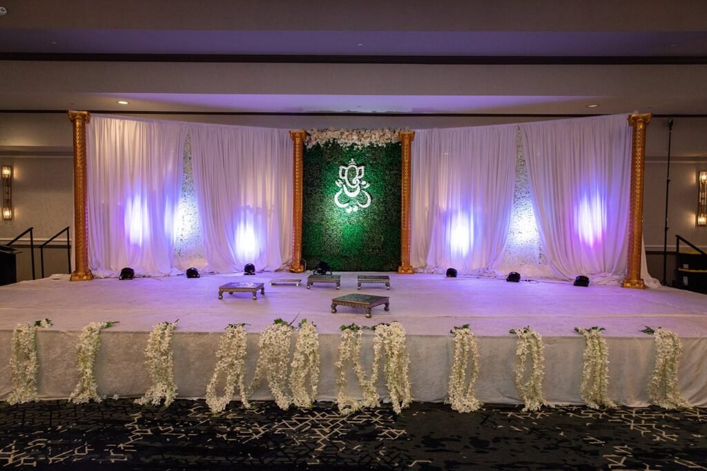 Floral Ganesha wedding stage design Pittsburgh — greenery wall with Ganesha motif, gold pillars, white drapes, luxury South Asian wedding decor