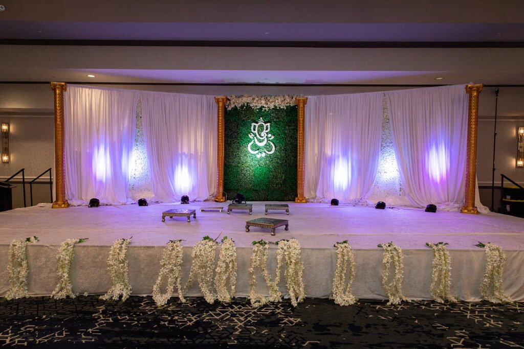 Floral Ganesha wedding stage design Pittsburgh — greenery wall with Ganesha motif, gold pillars, white drapes, luxury South Asian wedding decor